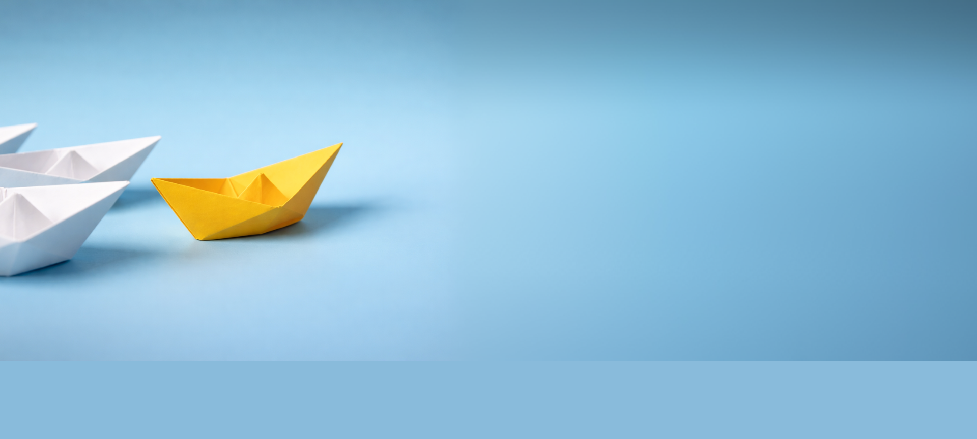 Ein gelbes Papierboot abgesetzt vor drei Weißen, mit viel freier Fläche. Das Bild betont Individualität und Führung.