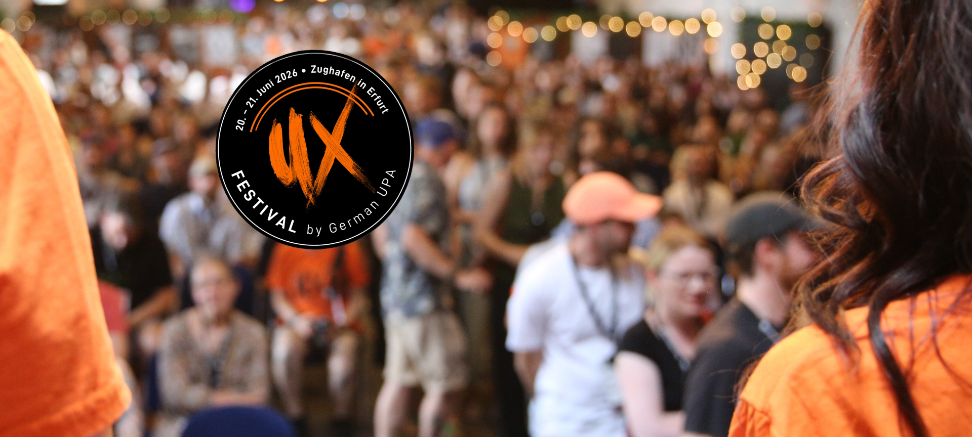 Unscharfe Menschenmenge auf dem UX Festival, gesehen von einer Bühne oder erhöhten Position. In der Mitte ein rundes Logo mit großen Buchstaben „UX“, umgeben von Text mit Datum und Ort.