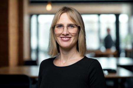 Franziska hat schulterlange blonde Haare. Sie trägt eine Brille und ein schwarzes Shirt. Sie lächelt breit.