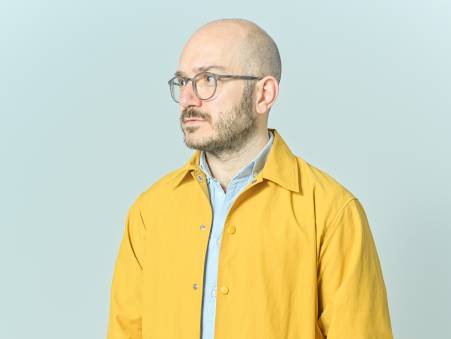 Jimmy Elias trägt ein gelbes Hemd und schaut abwesend an der Kamera vorbei.